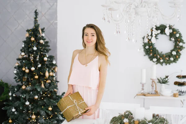 一个穿着漂亮粉色连衣裙的小女孩手里拿着一个金色礼品盒 手里拿着一个弓 放在客厅里的圣诞树后面 — 图库照片