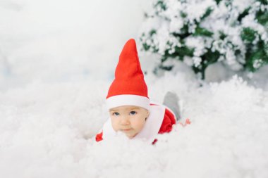 Noel Baba kostümü giymiş beş aylık bir bebek Noel ağacının altında yapay karın üzerinde yatıyor..