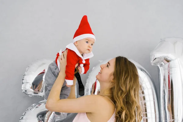 A young happy mother holds a baby in a Santa suit in her arms, against the background of the numbers 20. Christmas and New year 2020