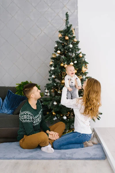 A young family sits on the floor in the living room near the Christmas tree, mom lifted up the baby in her arms, they are happy and smiling
