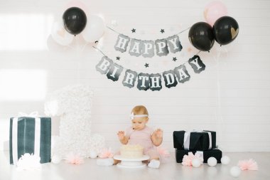 Pembe elbiseli bir bebek, hayatının ilk pastasını siyah beyaz fotoğraf bölgesinde mutlu bir doğum günü pankartıyla denedi.