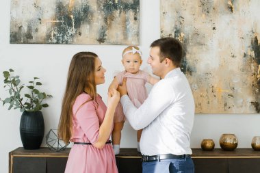 Oturma odasında ebeveynler ve bir yaşındaki kızları şifoniyerin yanında durup kızıma bakıyor ve ona gülümsüyor.