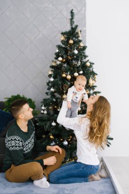Baba, anne ve bebek Noel ağacının yanında oturuyor. Annem bebeği kucağına aldı ve ona gülümsedi. Babam onlara baktı.