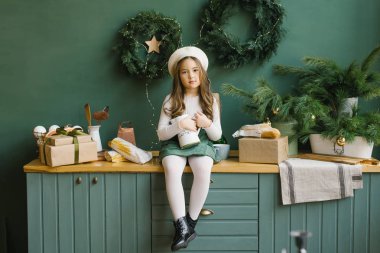 Elinde sürahi olan şık bir kız, Noel ve yeni yıl için zümrüt ve yeşil renklerle dekore edilmiş bir odada mutfak tezgahında oturuyor..