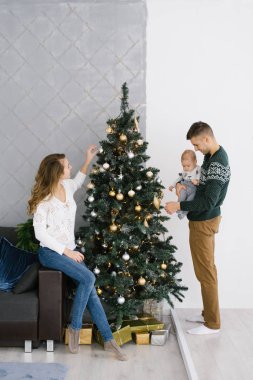Beyaz kazaklı ve kot pantolonlu genç bir anne Noel ağacının yanındaki koltukta oturur ve onu oyuncaklarla süsler. Kollarında bebeği olan baba yanında durur ve Noel ağacını süsler.