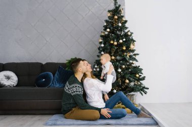 Bir anne ve baba, küçük bir çocuğu Noel ağacının yanında tutarlar. Evin içi Noel süsüsüyle süslenmiş. Aile tatili kavramı. Mutlu genç ve dost canlısı bir ailedir.
