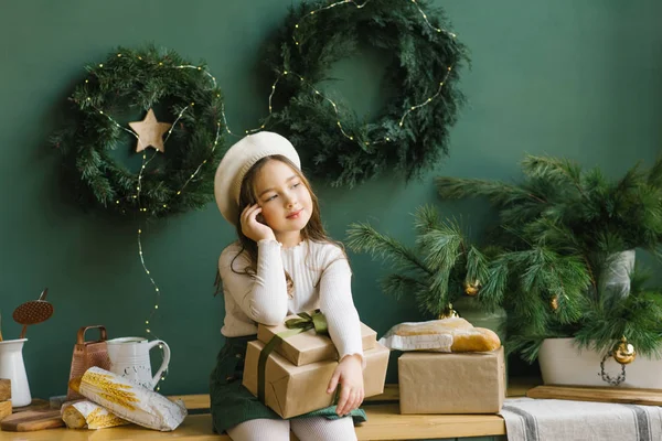 Cute little baby in a white beret with a Christmas gift box sitting on the countertop in the kitchen in green tones. She dreams and thinks about the holiday