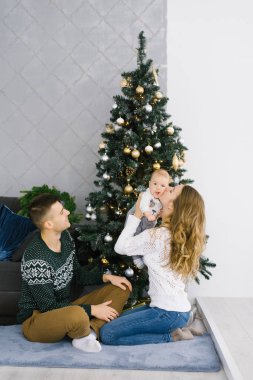 Mutlu Noeller ve yeni yıl. Genç bir aile tarafından kutlanıyor. Ebeveynler ve bebek Noel ağacının yanında oturuyor. Anne bebeği öpüyor.