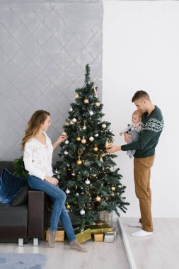 Noel ve yeni yıl için hazırlanıyor genç aile. Oturma odasında kucağında küçük bir çocukla Noel ağacını süsle.