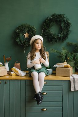 Beyaz bereli şık bir kız, Noel ve yeni yıl için süslenmiş şık bir mutfakta zümrüt ya da yeşille oturuyor. Duvarda Noel çelenkleri var. Kızın elinde beyaz bir sürahi var.