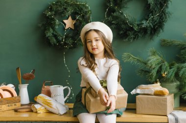 Beyaz bereli şık bir kız, Noel ve yeni yıl için süslenmiş şık bir mutfakta zümrüt ya da yeşille oturuyor. Duvarda Noel çelenkleri var. Noel hediyesi taşıyan bir kız