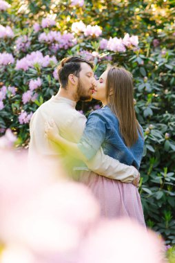 Aşık genç bir çift, çiçek açmış bir rhododendronun arka planında öpüşür. İlk randevu. Sevgililer Günü