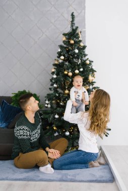 Mutlu Noeller ve genç ailenin oturma odasında yeni yıl kutlaması. Noel ağacının yanında yerde oturuyorlar, gülümsüyorlar ve mutlular.