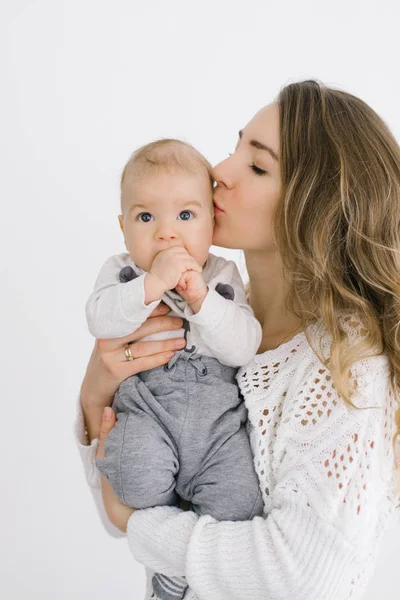 A young mother with blonde hair kisses her young son and holds him in her arms on a white background
