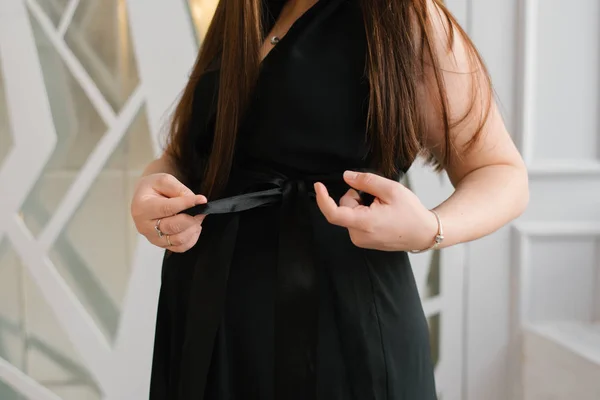 Pregnant woman ties a bow on her black dress on her stomach, close-up view of hands