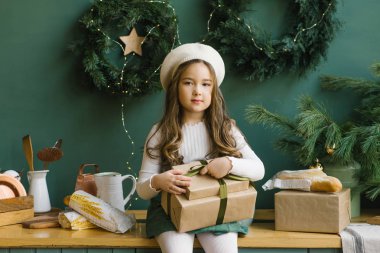 Beyaz bereli şirin bir kız elinde Noel hediyeleri ve zümrüt renkli mutfakta oturuyor. Yeni yıl tatili için süslenmiş.
