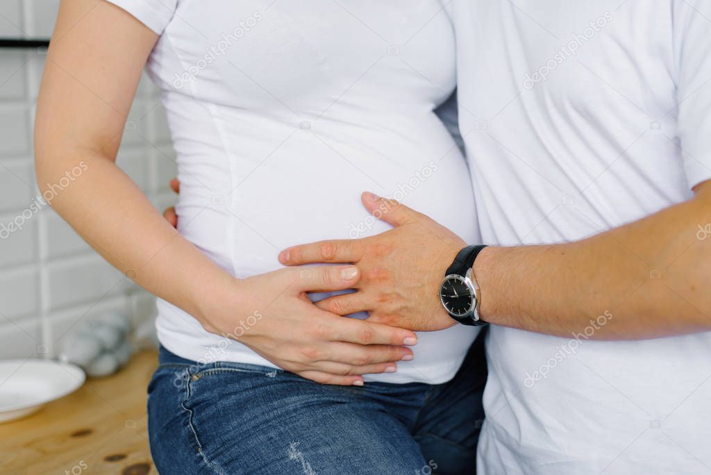 Expectant parents in white t-shirts and jeans. The husband holds his hand on the belly of a pregnant wife. Waiting for a baby in the family