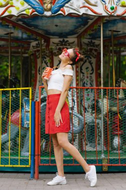 Kırmızı etekli parlak bir kız elinde kağıt bir bardak tutuyor, kırmızı güneş gözlüğü takıyor. O mutlu, gülümsüyor ve eğlence parkında dinleniyor.