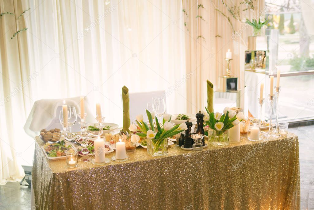 Decorated table of the bride and groom at a wedding Banquet with a tablecloth with gold sequins, white tulips in a glass vase and burning flowers