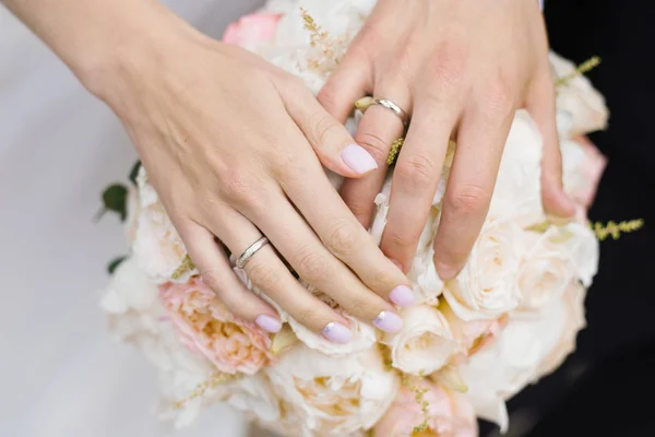 Handen van het pasgetrouwde echtpaar, de bruid en bruidegom, met trouwringen op een bruiloft boeket van witte en melk rozen en pioenen close-up. Bruidspaar — Stockfoto