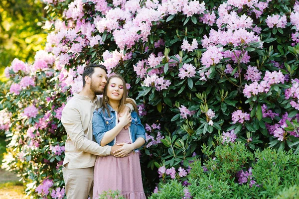 Uppmuntra känslor av kärlek. Ett par förälskade på en bakgrund av rhododendron blommor. Vacker flicka njuter av vårblommor. Killen kramar henne.. — Stockfoto