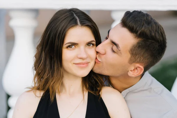 Bonito jovem casal apaixonado posando na rua. Um jovem beija uma mulher sorridente feliz na bochecha — Fotografia de Stock