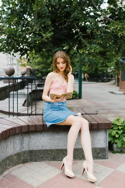 Jovem menina em roupas de verão com pernas longas sentado em um banco i — Fotografia de Stock