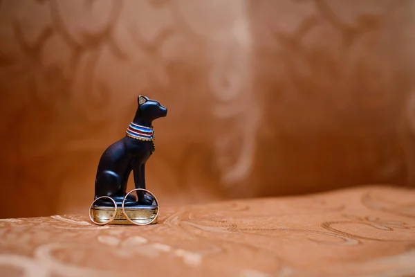 Gold wedding rings next to a statuette of an Egyptian black cat on a brown background