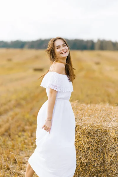 Una joven de pelo rubio con un vestido blanco en un campo con pilas de paja disfruta de un día de verano y sonríe — Foto de Stock