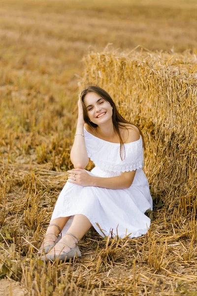 Una joven de pelo rubio con un vestido blanco en un campo con pilas de paja disfruta de un día de verano y sonríe — Foto de Stock