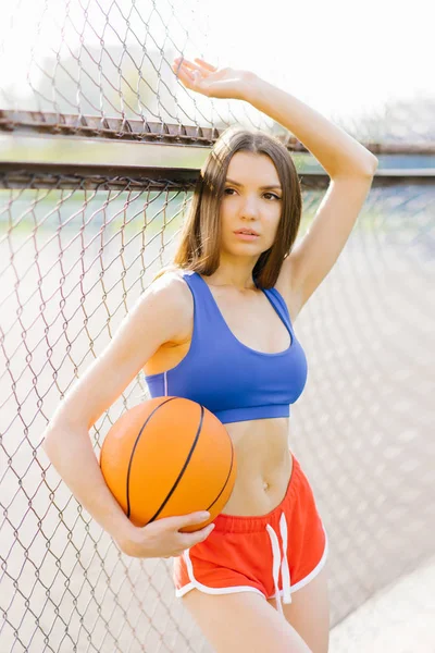 Girl in short red shorts and a blue top holds a basketball ball — Stockfoto