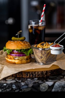 A front view of a cheeseburger and fries with a soft drink in behind, against a dark background.