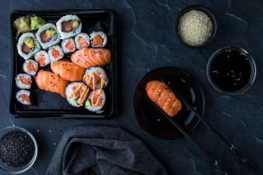 A top down view of assorted sushi against a dark background and surrounded by sesame seeds, soy sauce and chopsticks.