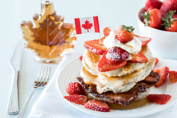 A close up of a stack of pancakes with a bottle of syrup and bowl of strawberries in behind. Canada Day concept. Stock Photo