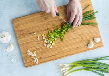 Tahta kesme tahtasındaki yeşil soğanı kesen ellerin yukarıdan görünüşü..