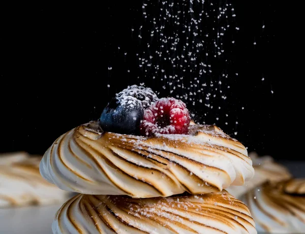 Un macro primer plano de merengues tostados con bayas y azúcar en polvo sobre un fondo negro . — Foto de Stock