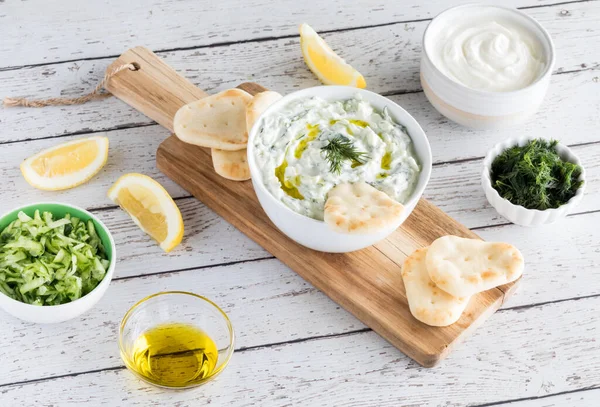 Vista de cima para baixo de uma tigela de molho tzatziki cercada por ingredientes usados para fazer o mergulho e picadas de pita para mergulhar . — Fotografia de Stock