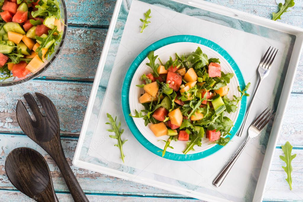 A top down view of a plate of watermelon and arugula salad in a tray with a bowl of the salad to the side.