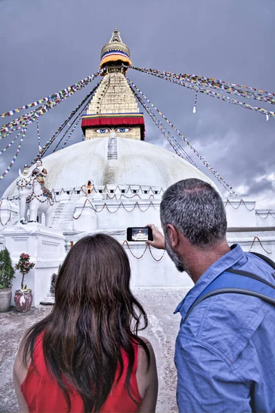 Western tourist couple. He with grey short and beard. Wearing a blue T-shirt and she with a large brown hair are taking photos and selfies with his smrtphone at the Boudhanath stupa