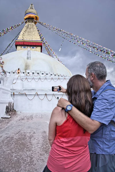 Western tourist couple. He with grey short and beard. Wearing a blue T-shirt and she with a large brown hair are taking photos and selfies with his smrtphone at the Boudhanath stupa