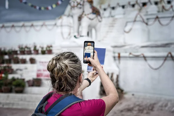 Sırtı önde ve sırt çantalı Batılı bir kadın. Saçları ve küpeleri toplanmış sarışın. Bir akıllı telefon ve Katmandu 'daki Boudhanath stupa' nın seyahat rehberiyle fotoğraf çekmek. Stu!