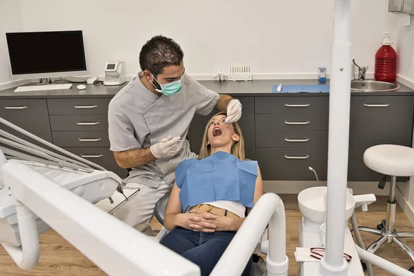 Dental doctor with blue mask checks the health of the mouth of his female patient lying in the armchair of a modern dental clinic. — Stock Photo, Image