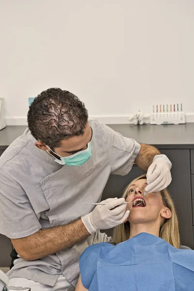 Dental doctor with blue mask checks the health of the mouth of his female patient lying in the armchair of a modern dental clinic. — Stock Photo, Image