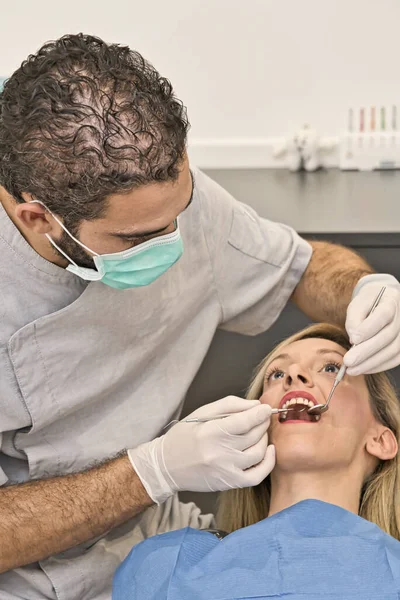 Mavi maskeli diş doktoru modern bir diş kliniğinin koltuğunda yatan kadın hastasının ağzını kontrol ediyor..