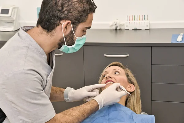 Dental doctor with blue mask checks the health of the mouth of his female patient lying in the armchair of a modern dental clinic. — Stock Photo, Image