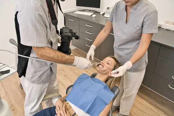 Dental doctor with blue mask checks the health of the mouth of his female patient lying in the armchair of a modern dental clinic. Photographing with a reflex the patient\'s teeth. Macro photography