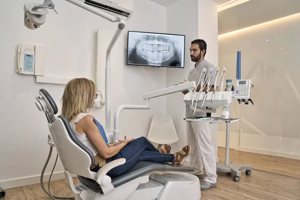 Black hair and beard dentist shows the client the x-ray of his mouth. Western client with blond hair was tumbling in the clinic chair. Modern dental clinic xray concept. — Stock Photo, Image