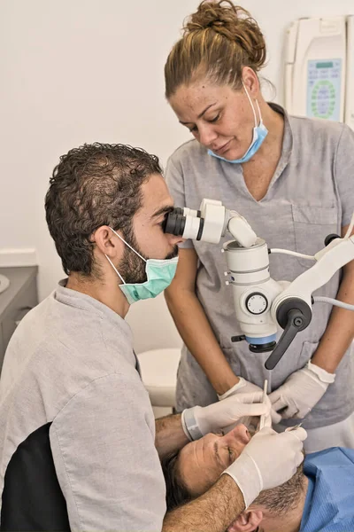 Dental doctor with microscope and blue mask next to his assistant checking his patient\'s dental health in a modern clinic