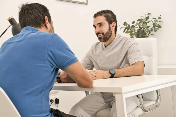 歯科医は、彼のオフィスで会い、テーブルに座って、現代の歯科クリニックで青いポロシャツを着ているクライアントに笑顔で出席します. ストックフォト