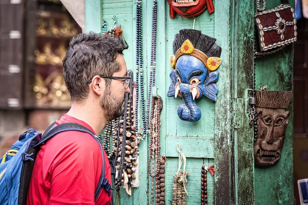 Asyalı, kısa siyah sakallı ve gözlüklü Batılı çocuk Nepal, Asya 'da bir dükkanda asılı duran turkuaz ahşap kapıya asılı maske ve hediyelik eşyalara bakıyor..